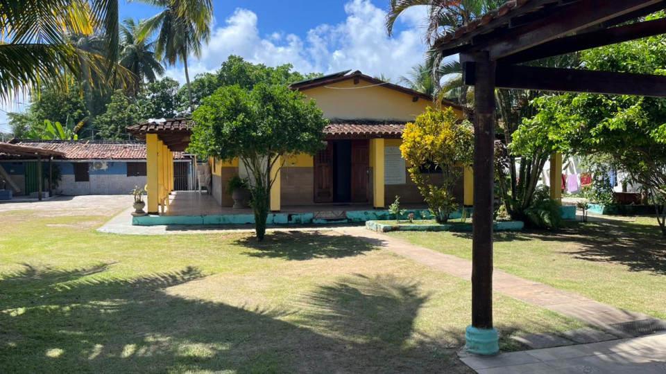 Fachada principal e infraestrutura da clínica de recuperação feminina em Ilhéus, Bahia, destacando uma casa amarela com varanda cercada por amplo gramado verde, árvores e ambiente de natureza tranquilo para o tratamento.